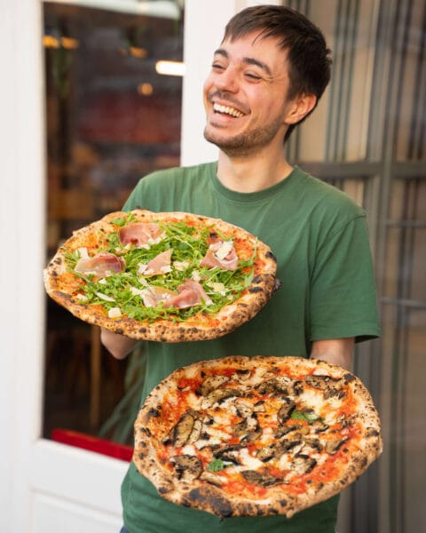 Guy holding Neapolitan pizzas topped with tomatoes, cheese, mushrooms, rocket, olives, prosciutto and parmesan shavings